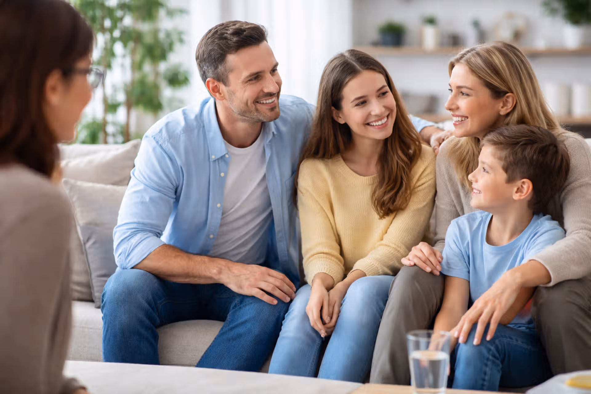 Assis sur un canapé beige, une famille est souriante. L'homme et la femme se regardent, entre eux se trouve une jeune fille qui regarde sa mère. Un jeune garçon est assis entre les jambes de sa mère et il regarde son père. En face de cette famille, et à gauche de l'image, une jeune psychothérapeute, de profil, brune, portant des lunettes, les regarde et sourit.